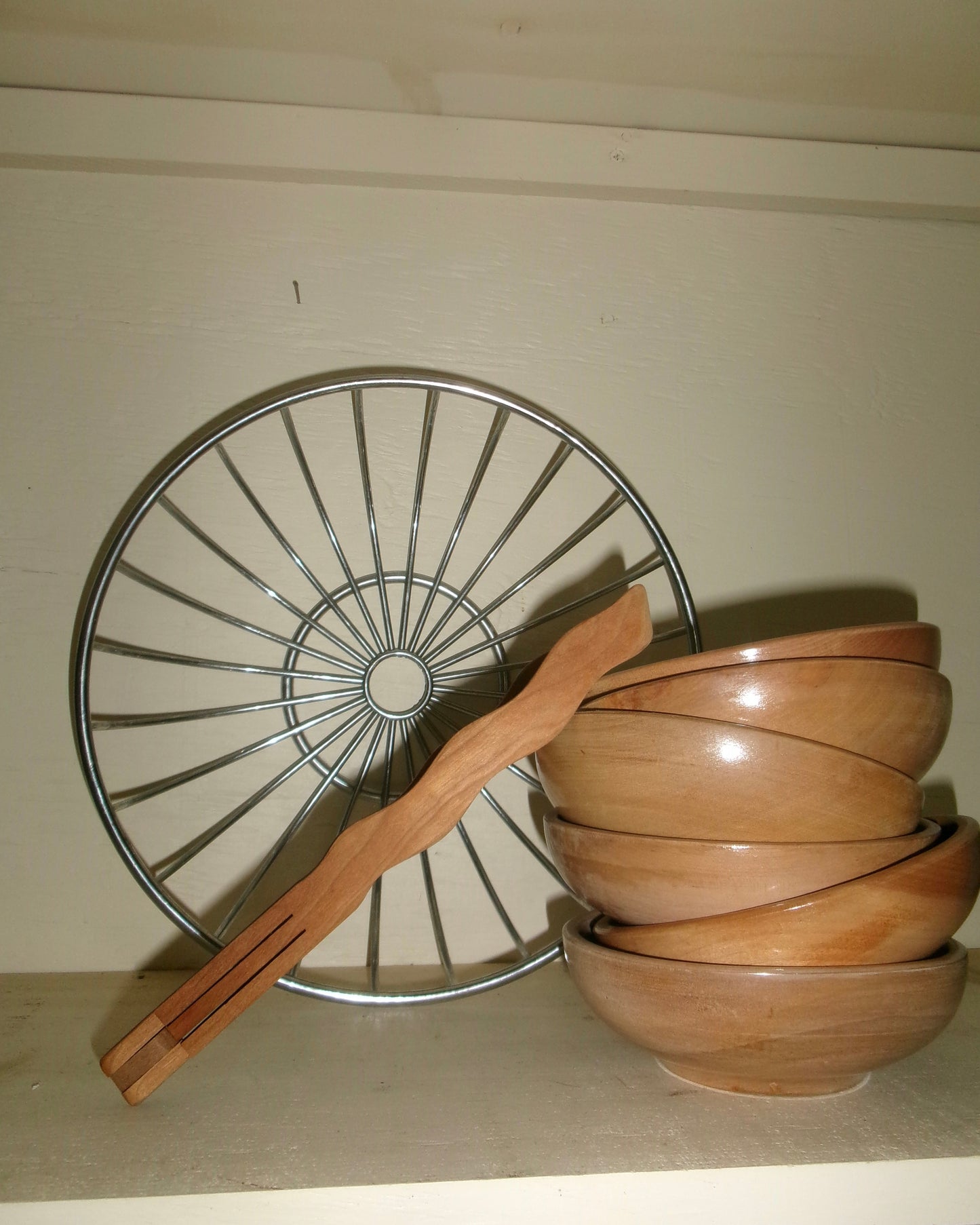 Wavy Wooden Tongs leaned against a stack of wooden bowls on a shelf with a large chrome bowl in the background.