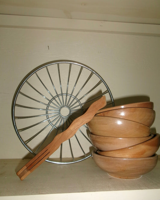 Wavy Wooden Tongs leaned against a stack of wooden bowls on a shelf with a large chrome bowl in the background.