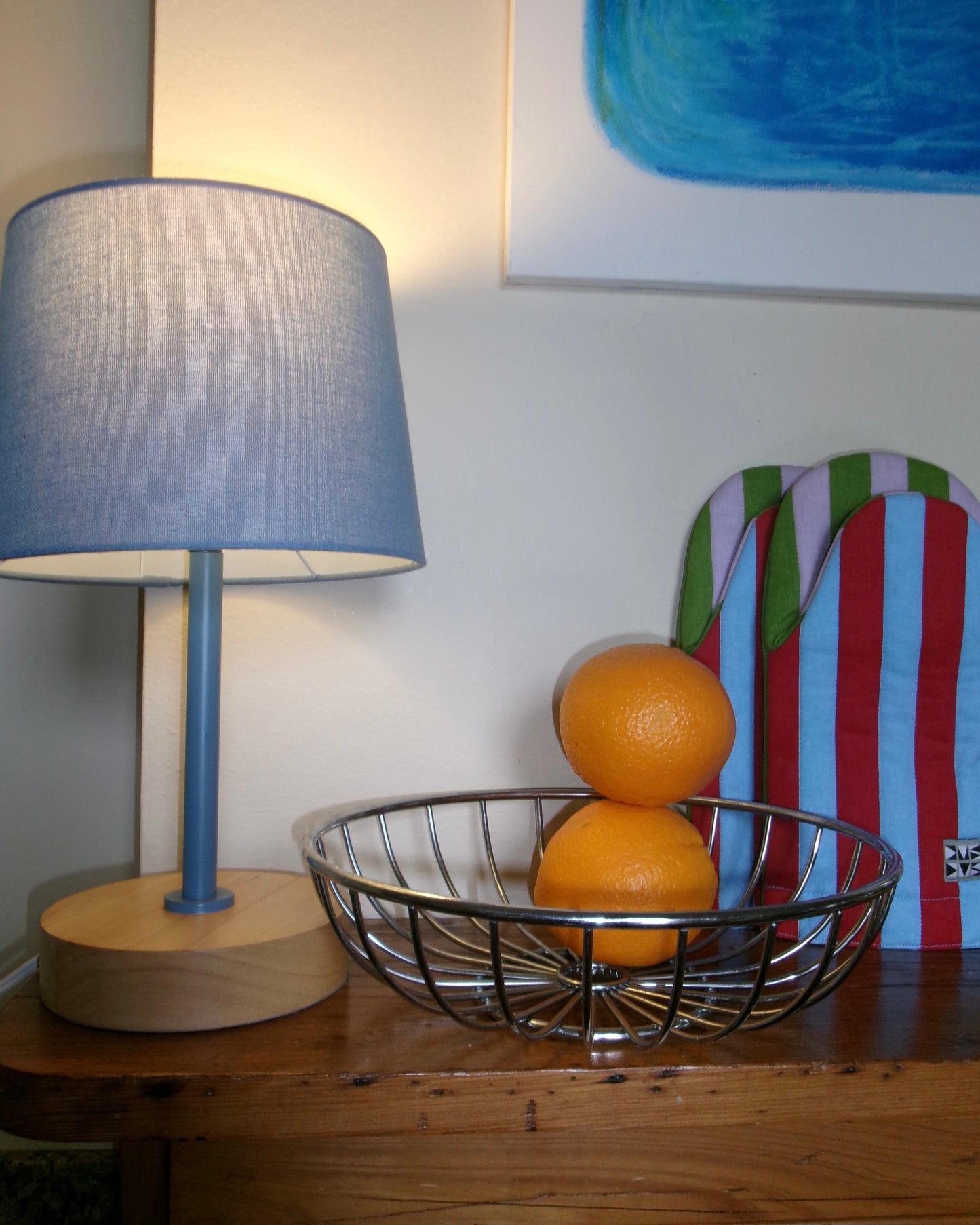 2 oranges in a chrome bowl next to a blue lamp and striped pot holders in a colorful, playful kitchen