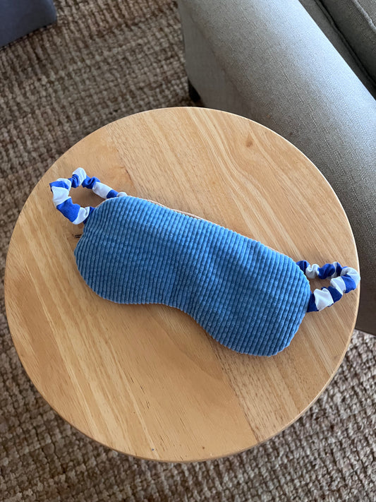 Blue corduroy sleep mask with striped elastic band displayed on a wooden surface